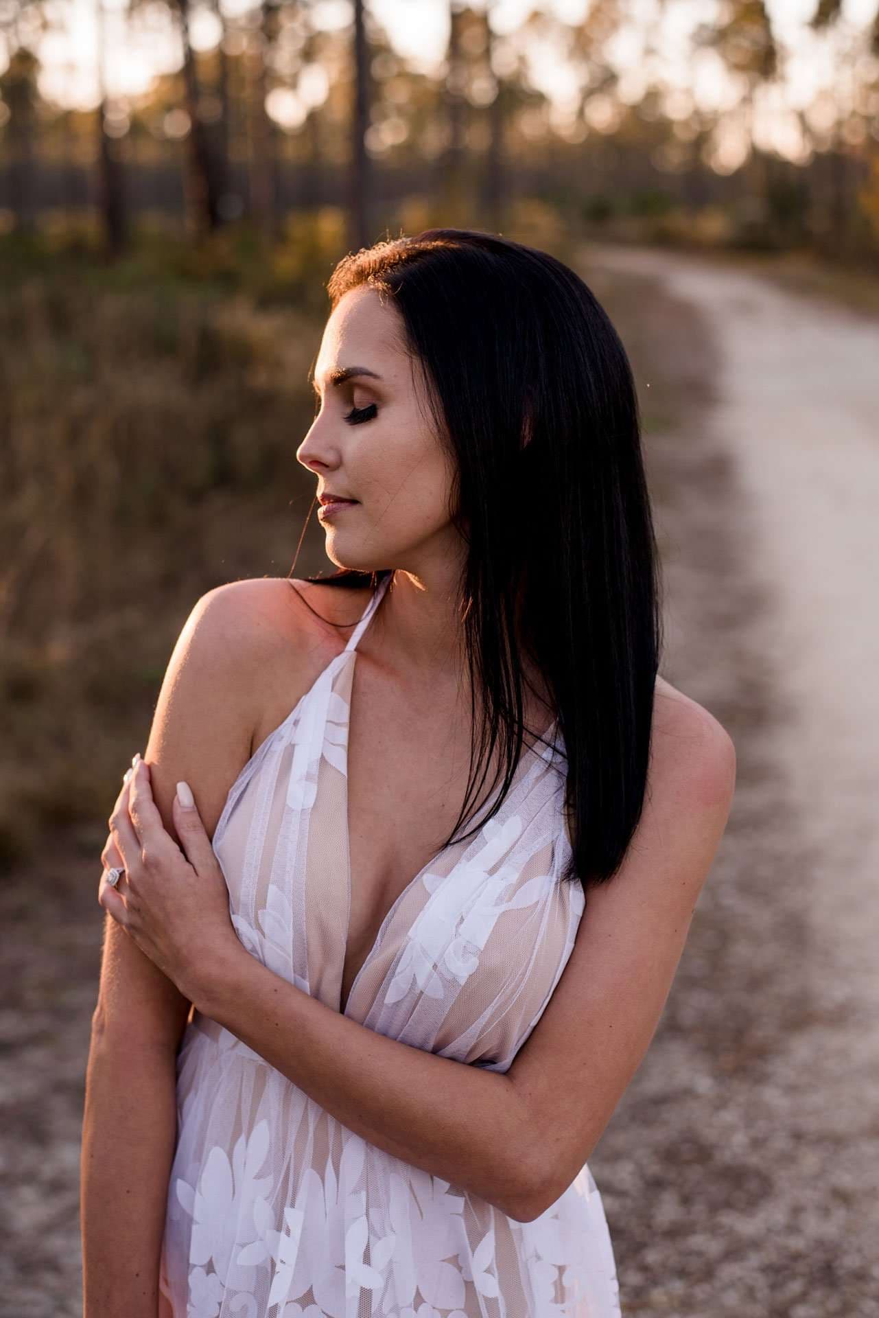 Woman with long dark hair and eyes closed, wearing a white lace halter dress, on a sandy path at sunset